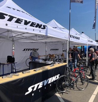 Weißer 3x3 m Eventzelt mit Stevens-Bikes-Logo für eine Radsportveranstaltung mit schwarzen halbhohen Seitenwänden und Theken