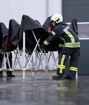 eutscher Feuerwehrmann in Uniform baut an einem regnerischen Tag einen schwarzen 3x3 m Faltpavillon von Ecotent auf.