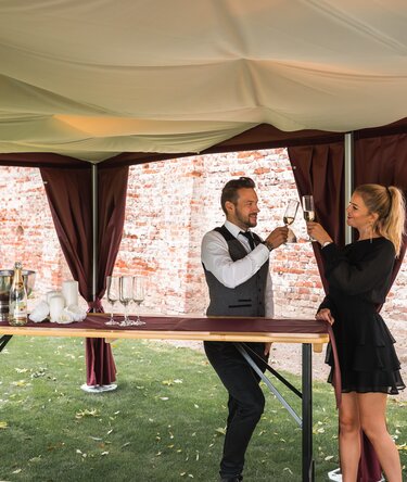 Unter dem eleganten Faltpavillon mit Himmel stehen 2 Personen mit einem Glas Prosecco in der Hand. 