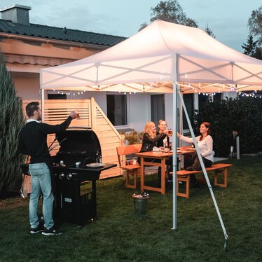 Freunde feiern unter dem Gartenzelt eine Grill- und Gartenparty. Das Gartenzelt ist feierlich dekoriert mit Lichterketten.