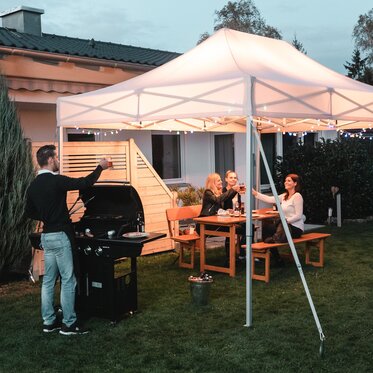 Freunde feiern unter dem Gartenzelt eine Grill- und Gartenparty. Das Gartenzelt ist feierlich dekoriert mit Lichterketten.