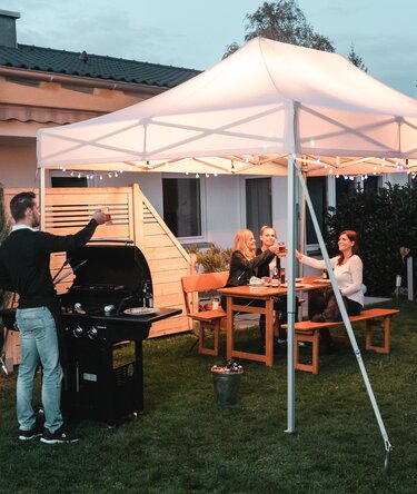 Freunde feiern unter dem Gartenzelt eine Grill- und Gartenparty. Das Gartenzelt ist feierlich dekoriert mit Lichterketten.