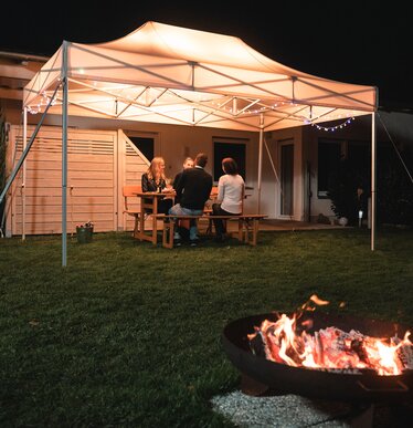 Das 4,5x3 m Gartenzelt steht im Dunkeln im Garten. Darunter sitzen die 4 Gäste. Vor dem Zelt ist noch eine Feuerstelle.