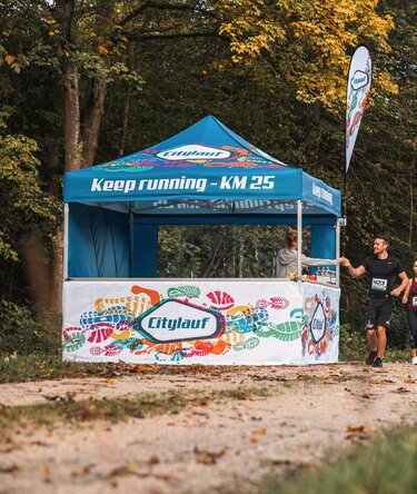 Blauer 3x3m Eventzelt mit individuellem Citylauf-Druck, mit individueller Fahne, geschlossener Seitenwand und halbhoher Seitenwand mit Theke als Verpflegungsstand bei einem Stadtlauf