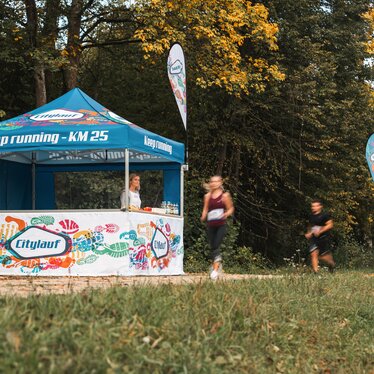 Das blau-weiße Eventzelt dient als Versorgungsstation bei einem Marathon. 2 Läufer laufen gerade am Eventzelt vorbei.