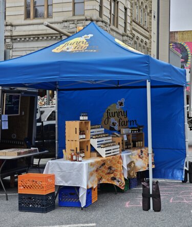 Auf einem Markt steht ein blauer, bedruckter Ecotent Faltpavillon Serie E1 der Größe 3 x 3 Meter mit Seitenwand von Funny Farm. 