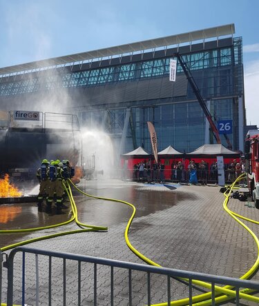 Feuerwehrteam bei einer Brandbekämpfungs-Demonstration im Freien auf einer Messe, mit Wasserstrahl, Einsatzfahrzeugen und Einsatzzelten im Hintergrund.